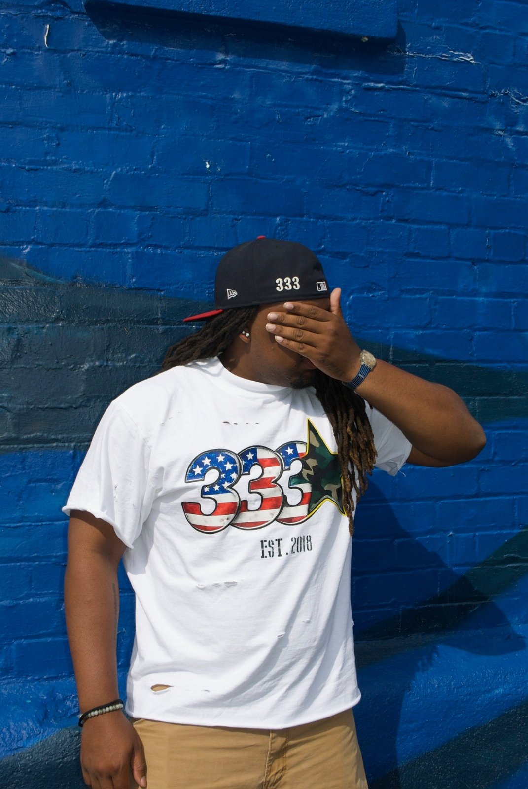 Portrait in the white 333 Est. 2018 tee and 333 New Era cap against a cobalt wall