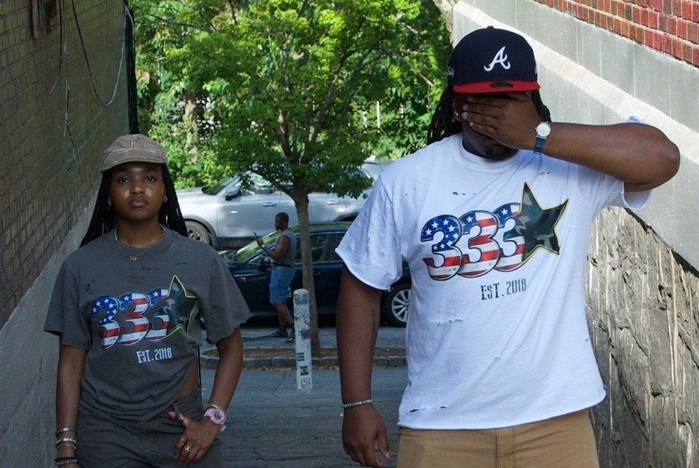 Two wearers in 333 Est. 2018 tees — white and grey — walking a shaded Brooklyn alley